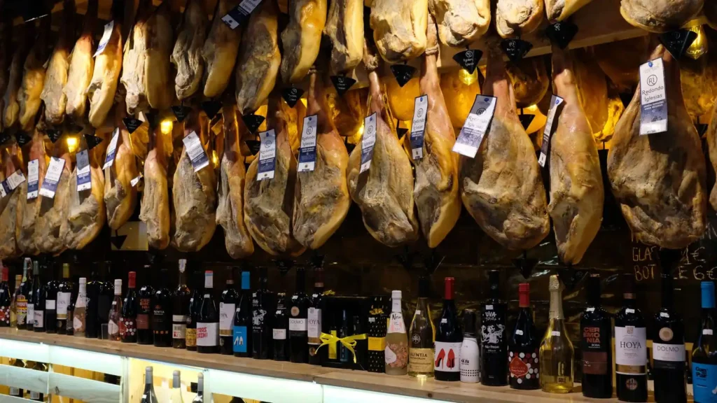 Jamones colgados con sus etiquetas sobre una fila de botellas de vino español en una tienda de Barcelona